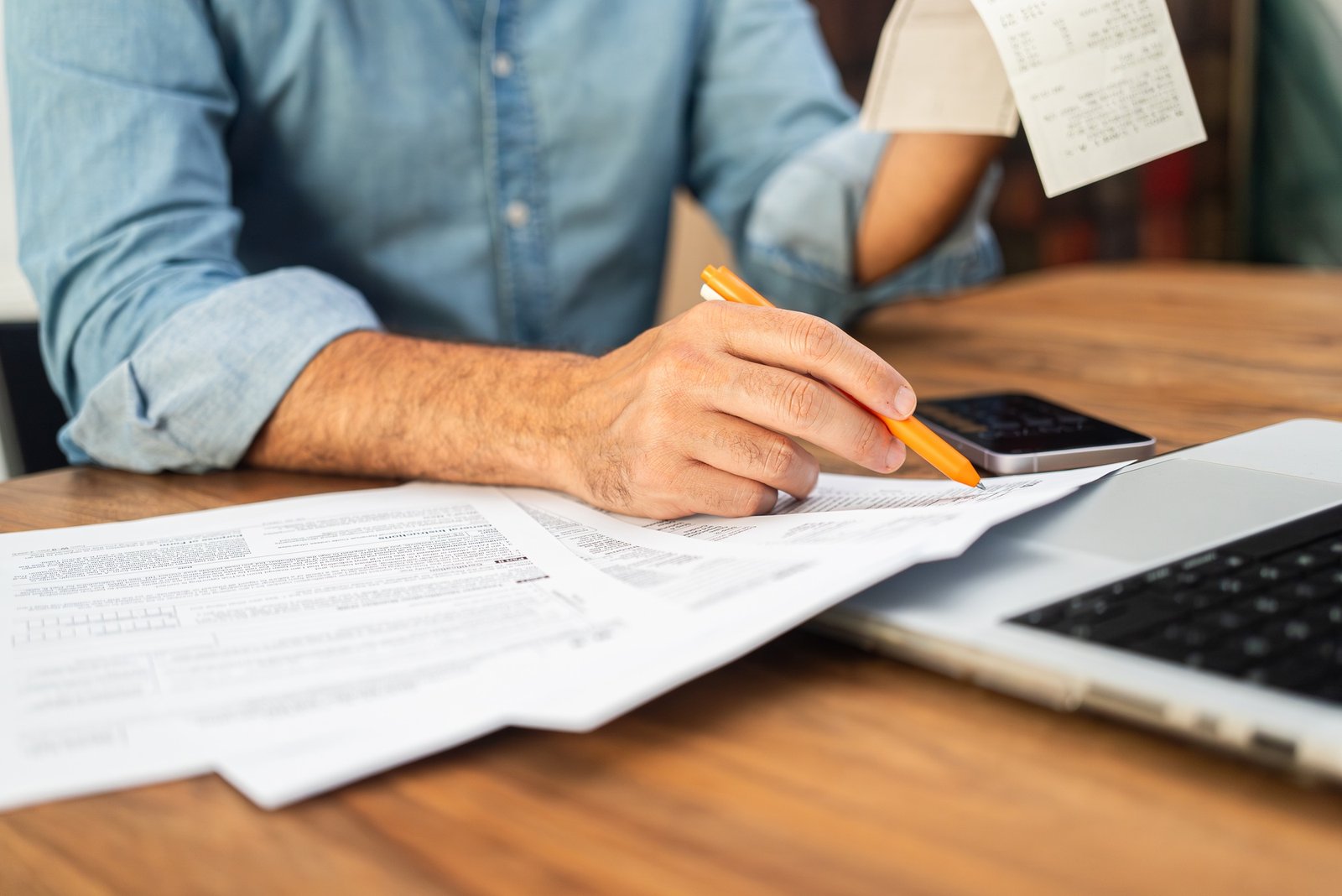 Man in a denim shirt calculating taxes with receipts and forms on a wooden desk.