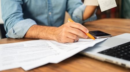 Man in a denim shirt calculating taxes with receipts and forms on a wooden desk.